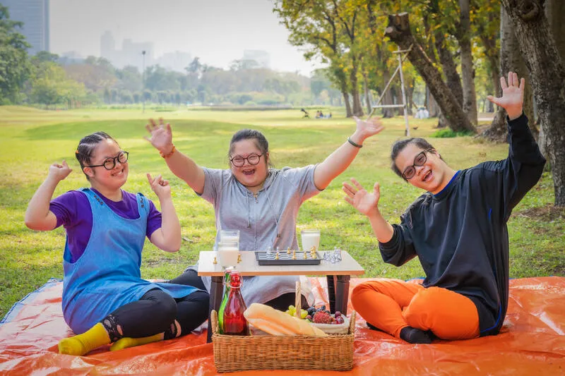Three friends smiling and waving at a picnic in a park